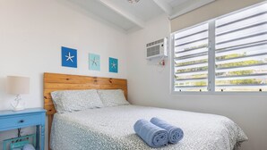 Ein Schlafzimmer mit einem Holz-Kopfteil, einem Bett mit einem gemusterten Überwurf, einem Nachttisch mit einer Lampe und einem Fenster mit horizontalen Jalousien.
