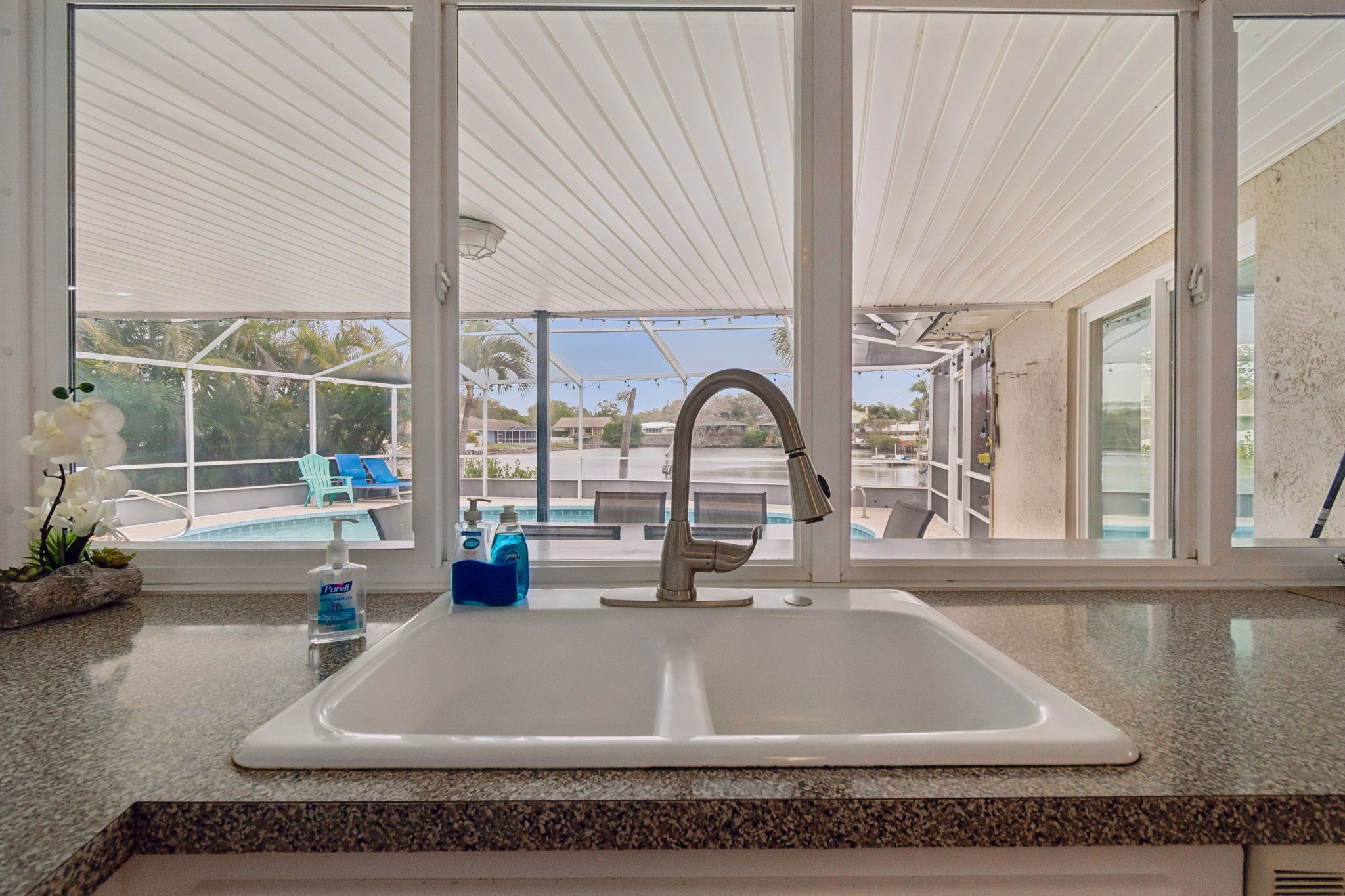 A kitchen with a white sink, granite countertop, and a view of a pool and palm trees.