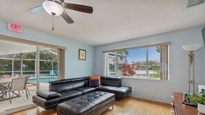 A modern living room with a sectional sofa, a ceiling fan, a large window with a view, and an exit sign. - Waterfront Pool Home - 12PPL 4BR (Clearwater)