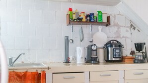 Une cuisine avec un mur recouvert de carreaux blancs, une étagère en bois où sont rangés divers ustensiles de cuisine, une machine à café, un cuiseur à riz et une bouilloire.