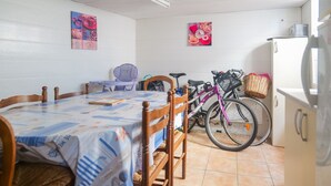 Un coin repas avec une table recouverte d’une nappe, des chaises en bois, un vélo et un réfrigérateur.