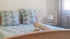 Un lit bien fait, recouvert d’une couvre-lit à motifs, avec deux oreillers ornés de motifs de feuilles, une serviette enroulée, une lampe de chevet et une fenêtre avec des rideaux.
