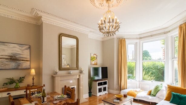A well-lit living room with a fireplace, a dining area with wooden furniture, a television, and a large window with curtains. - Cosy Flat in the Heart of Jesmond - Pass the Keys (Newcastle)