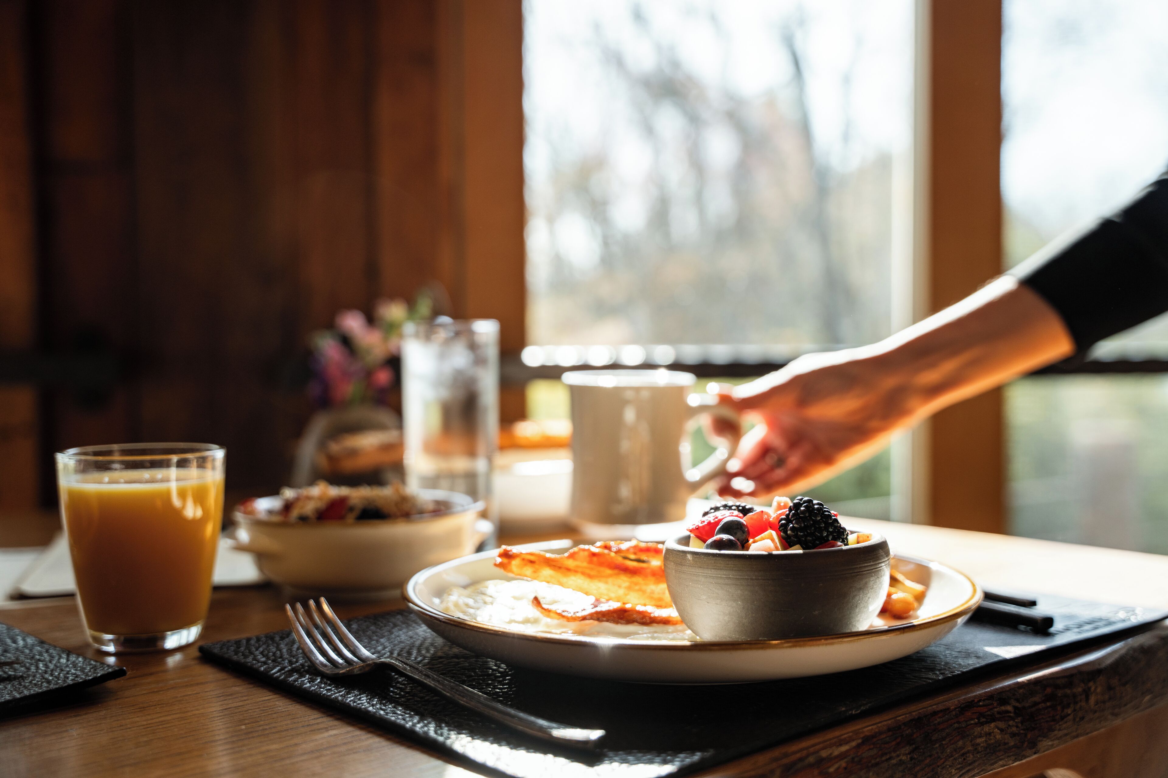 A breakfast setting with a plate of pancakes, a bowl of fruit, and a glass of orange juice on a wooden table.