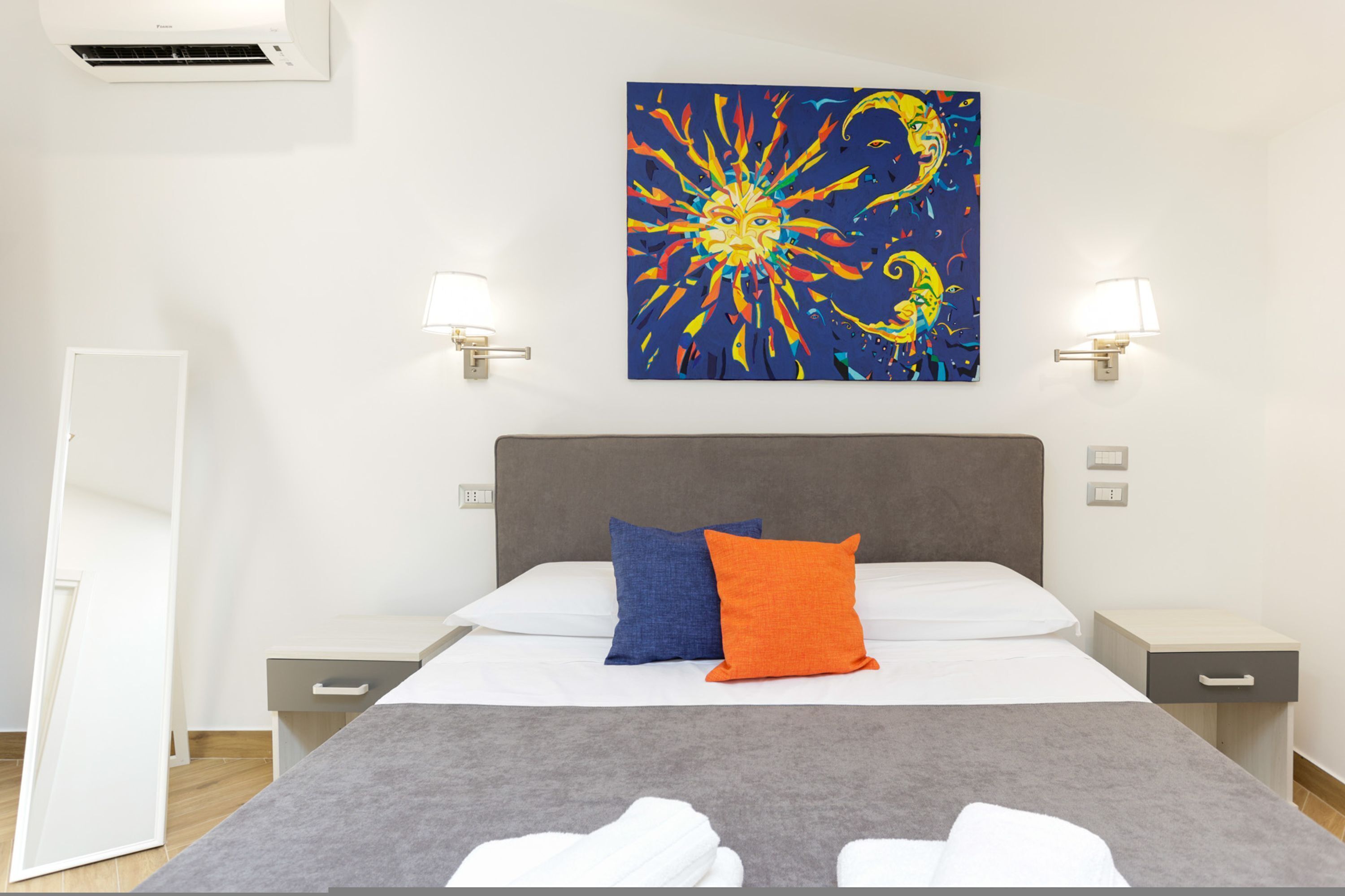Une chambre d’hôtel avec un lit, deux tables de chevet, une petite table, un climatiseur fixé au mur et un tableau coloré au-dessus du lit.