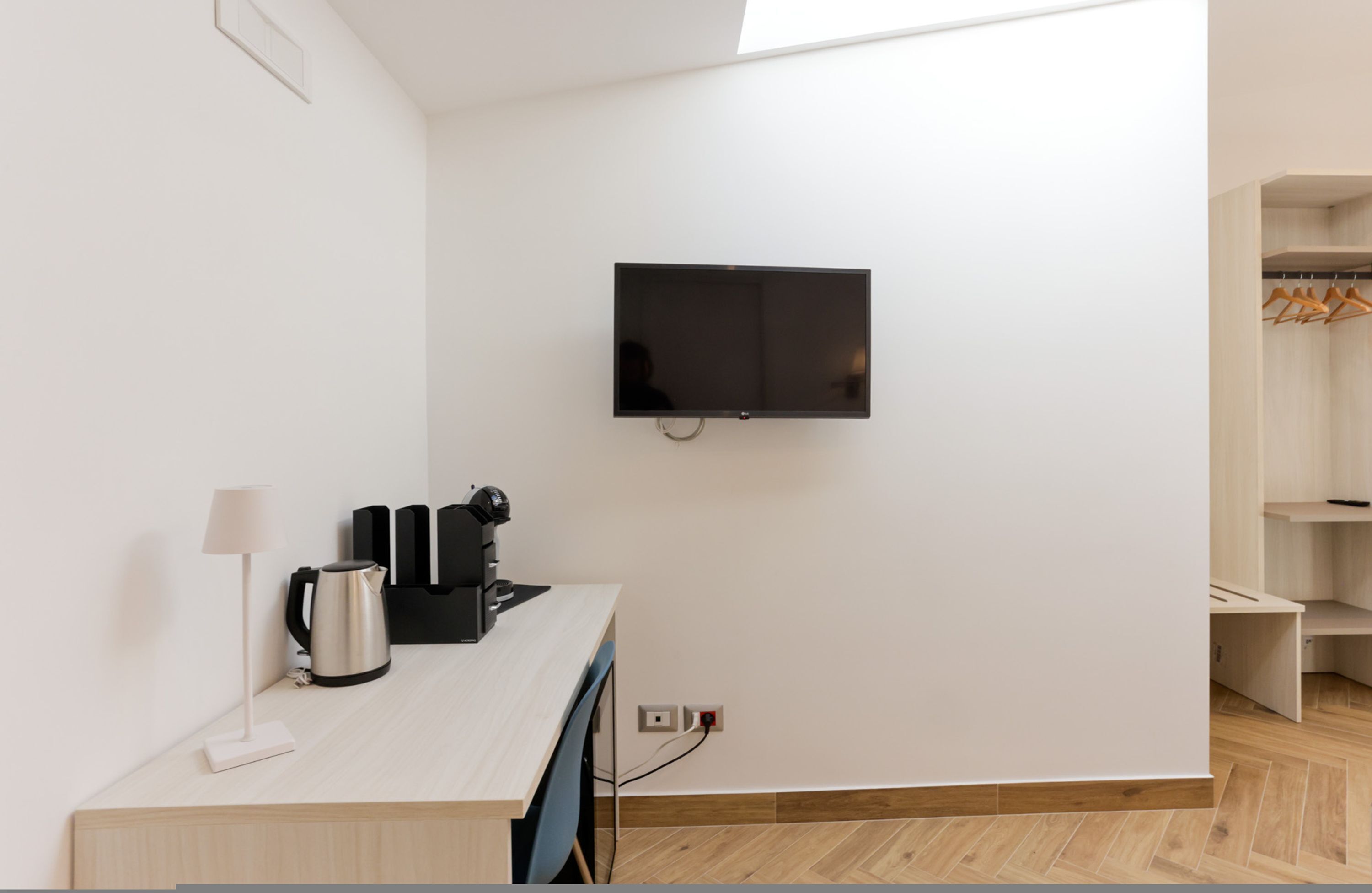 A modern room with a flat-screen TV mounted on the wall, a wooden desk with a lamp, a coffee maker, and a power outlet.
