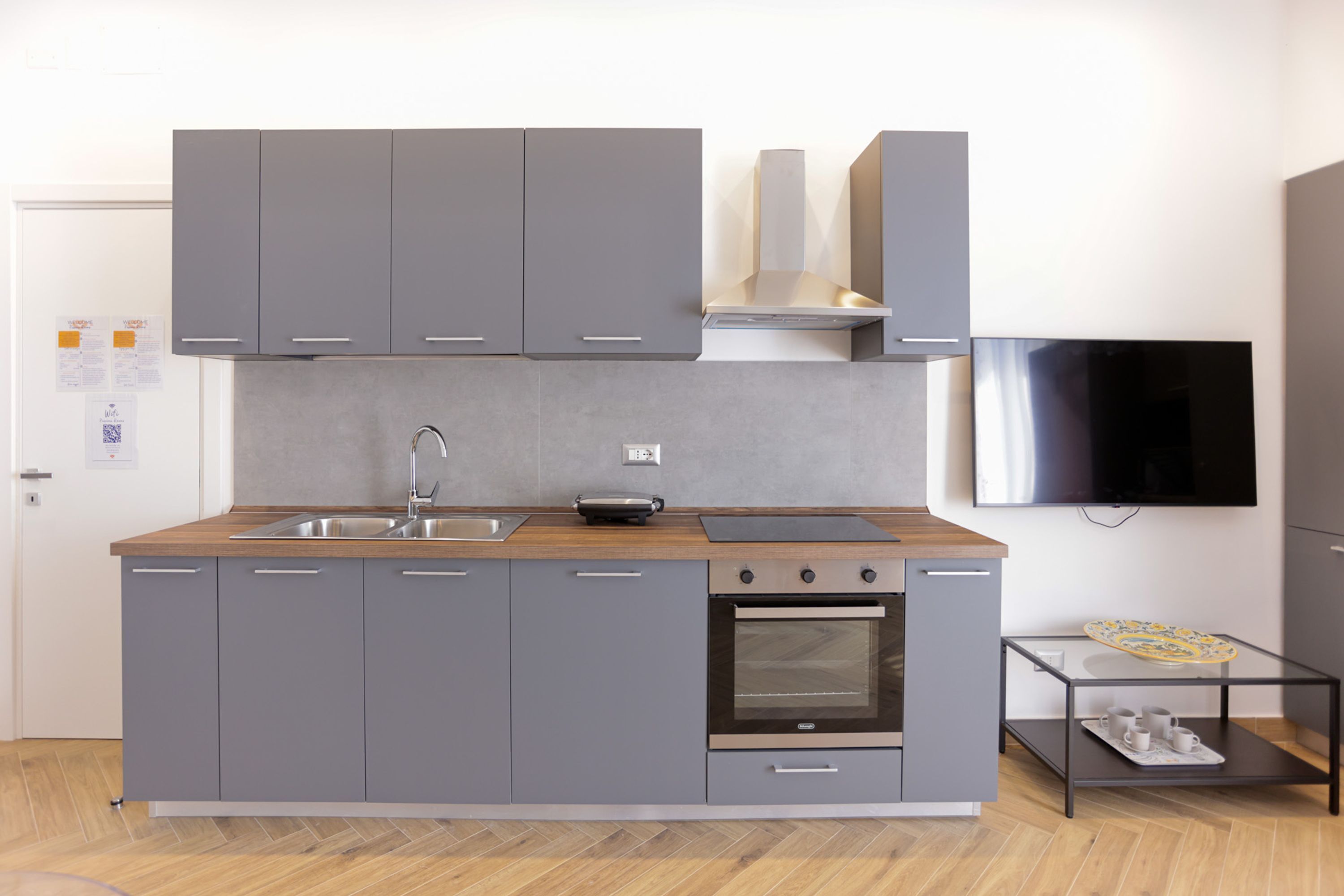Eine moderne Küche mit grauen Schränken, einer Holz Arbeitsplatte, einem eingebauten Ofen und einem an der Wand montierten Flachbildfernseher.