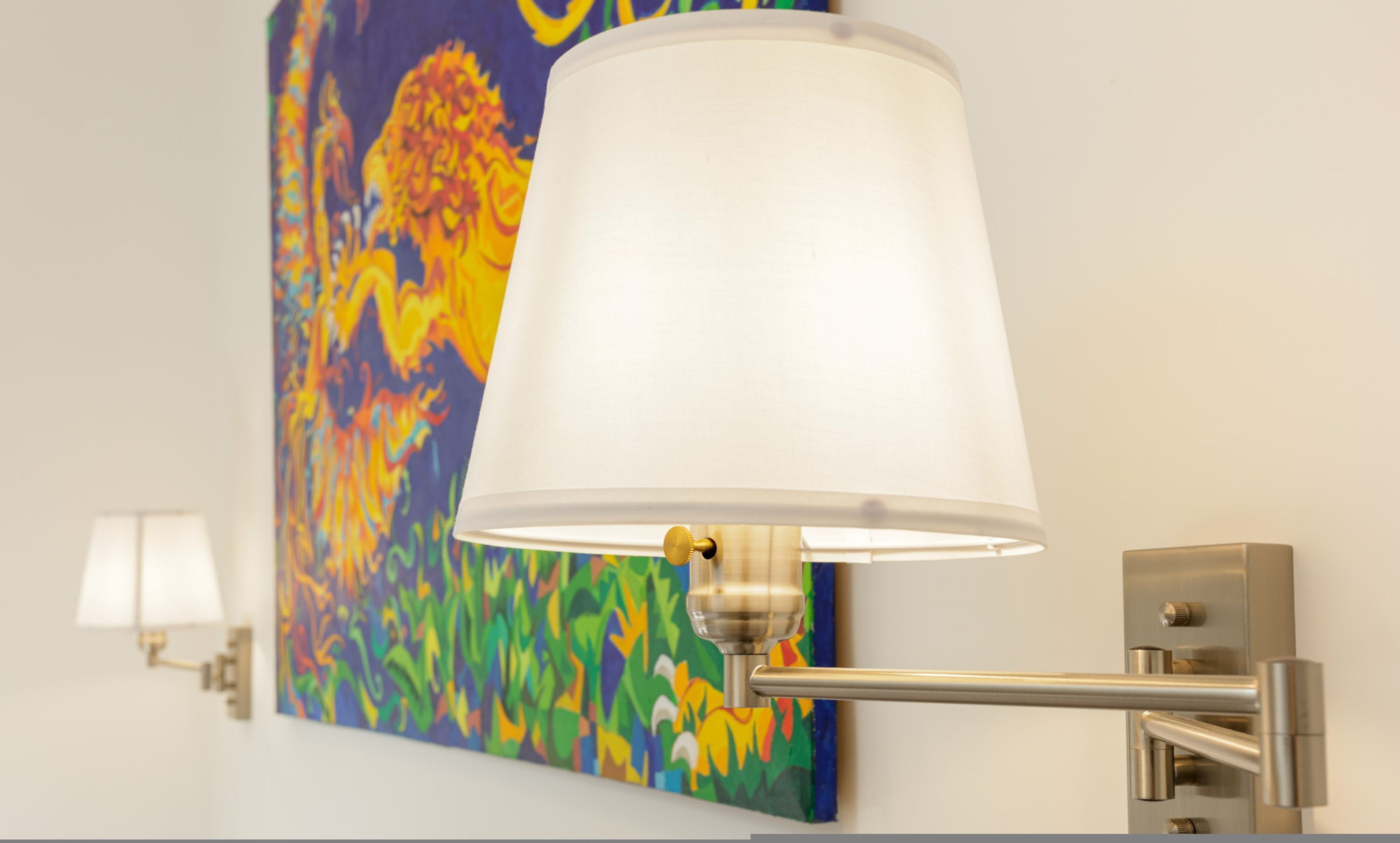 Une lampe murale dotée d’un abat-jour blanc et d’un pied métallique, placée devant une peinture abstraite aux couleurs vives.