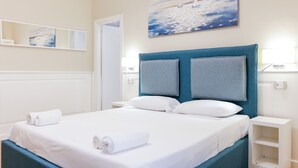 Een net opgemaakt bed met witte lakens en twee kussens, een blauw hoofdbord, een schilderij van zeilboten aan de muur en een witte nachtkastje met een lamp.