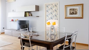 A modern living room with a glass dining table, white chairs, a flat-screen TV, and a sofa with yellow cushions. - Riviera di Chiaia 207 (Naples)