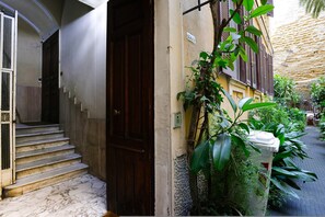 Un bâtiment doté d’un escalier en marbre menant à une porte en bois, d’un petit jardin avec des plantes et d’un mur à la texture particulière.