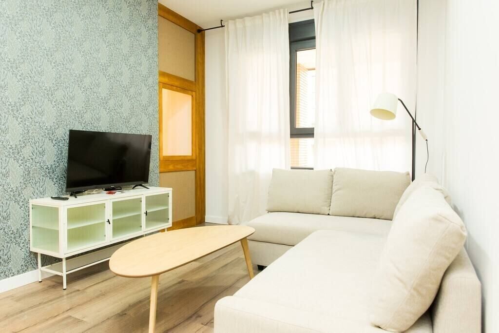 Een woonkamer met een televisie op een wit meubelstuk, een beige bank en een kleine tafel.