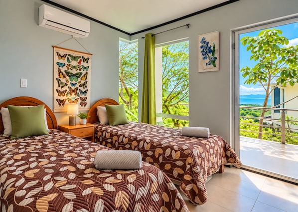 A hotel room with two beds, a window with a view of greenery and the sea, and a wall-mounted air conditioner.