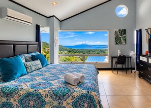 A bedroom with a large bed, a view of the sea, a desk, and a chair.