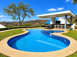 A swimming pool with a curved edge, a covered outdoor seating area, and a view of hills in the background.