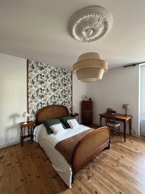 A bedroom with a wooden bed, a wall-mounted ceiling light, a small wooden table with a lamp, and a chair. - Située au Coeur des Vignobles du Médoc, Près de Bordeaux et des Plages Océanes (Saint-Estèphe)