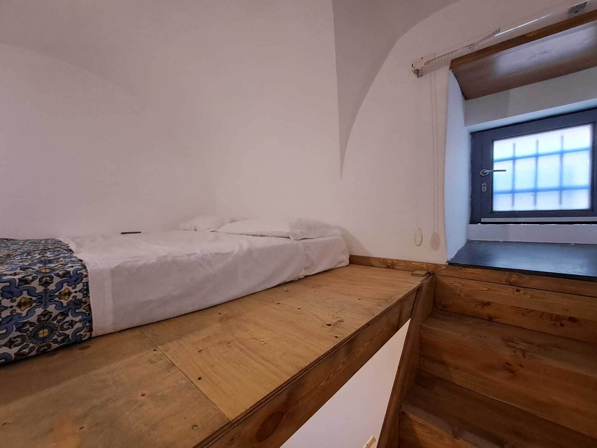 Une petite pièce, de taille modeste, comprenant un lit simple, un banc en bois et une fenêtre donnant sur un paysage agréable.