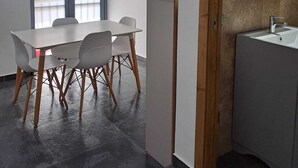 Een moderne kamer met een eettafel, stoelen, een groot raam en een badkamer met spiegel en wastafel.