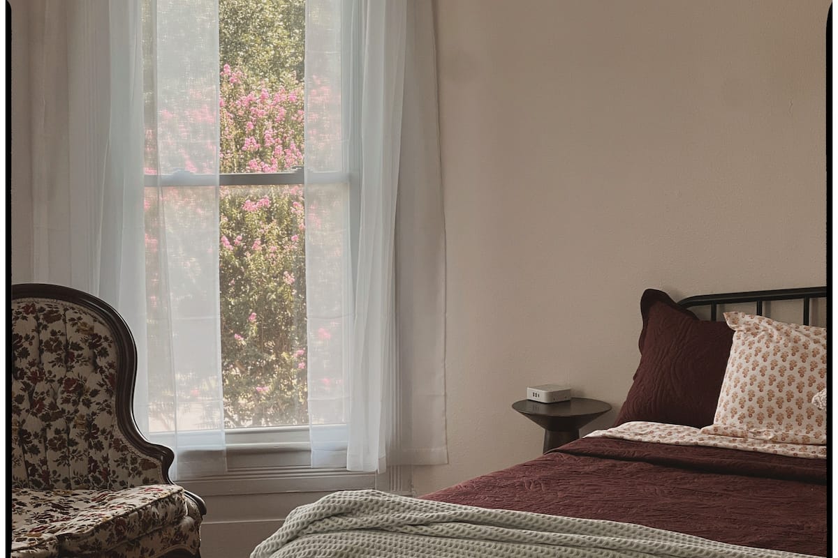 Quarto com cama, cadeira, mesa de cabeceira e janela com cortinas leves.