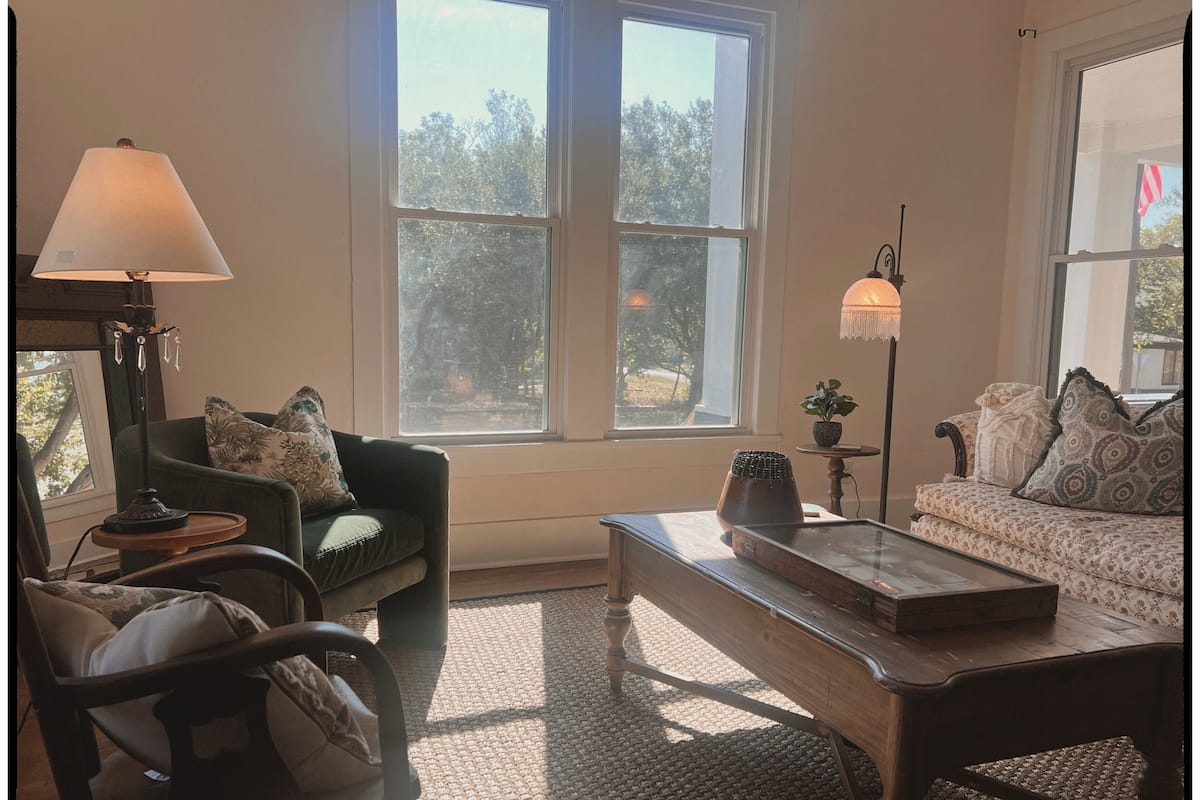 Uma sala de estar acolhedora com sofá, poltrona e mesa de centro, janelas amplas com vista para árvores e uma luminária sobre uma mesinha lateral.