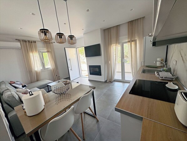 A modern kitchen with a built-in oven, a dining area with a glass table and chairs, and a living area with a sofa and a flat-screen TV.