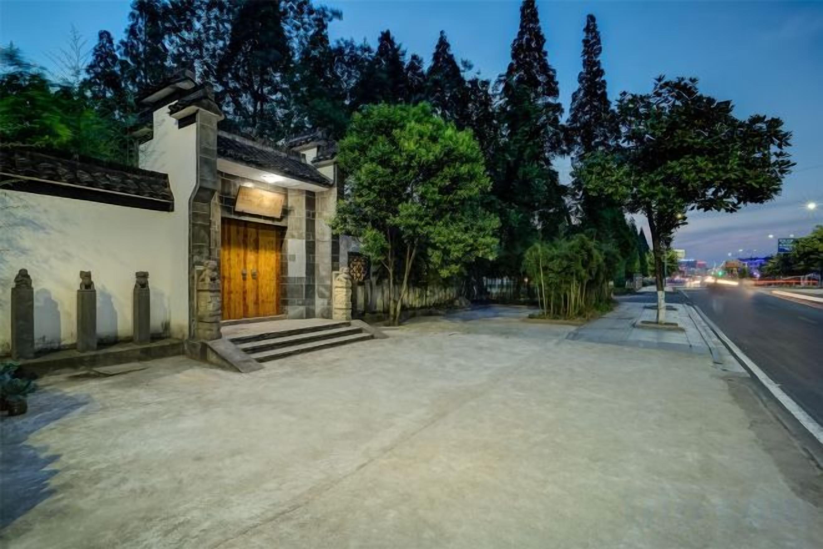 A building with a wooden door and steps leading to the entrance, surrounded by trees and a paved area.