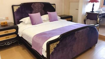 A bed with a purple headboard and white bedding, a nightstand with a lamp, and a wooden floor.