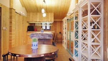 A wooden interior with a round dining table, chairs, a kitchen area, and a white shelving unit.