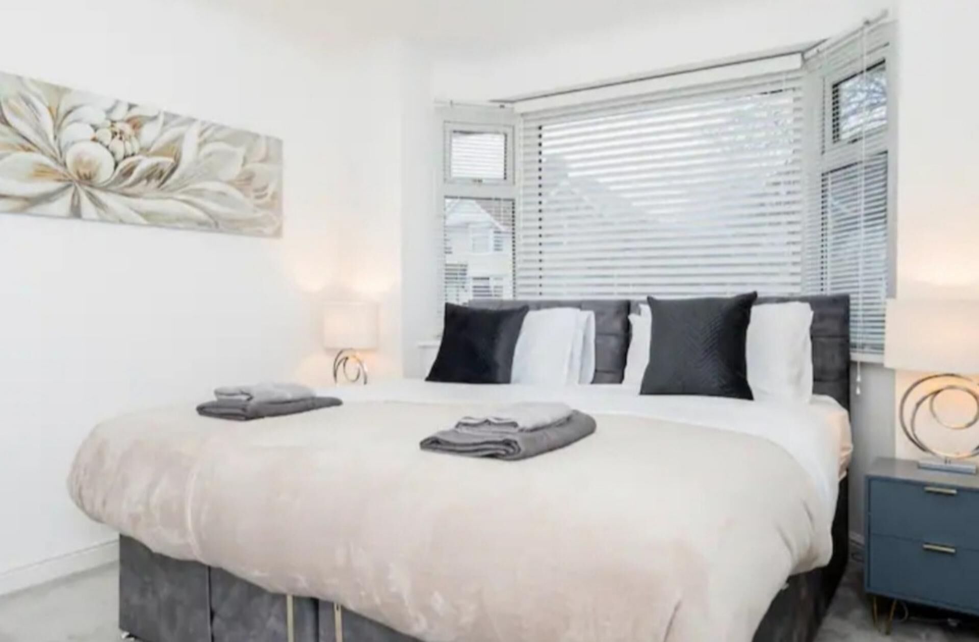 A modern bedroom with a large bed, white bedding, a side table, and a wall-mounted artwork.