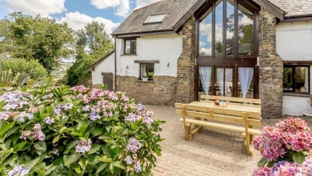 Une maison à deux étages avec une façade en pierre, de grandes fenêtres et un banc en bois dans le jardin.