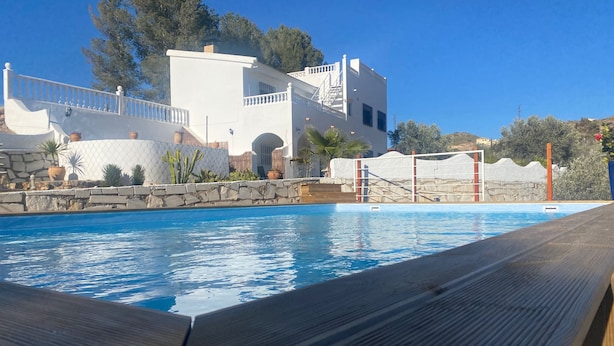 A swimming pool with a wooden deck in the foreground, a white building with a stone wall and potted plants in the background, and a clear blue sky. - 3beds Villa w/ Pool - Near Almeria Beaches (Cantoria)