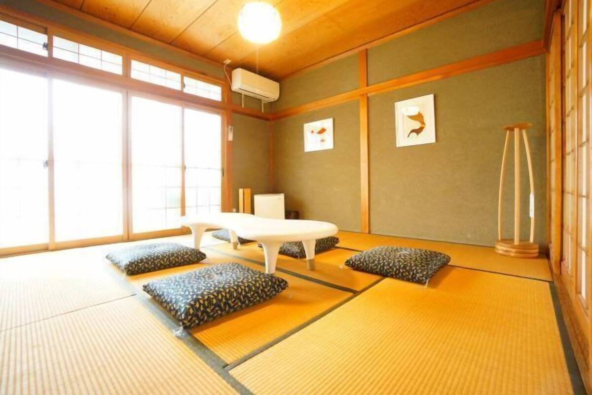 A tatami mat room with a low table and cushions, large windows, and a traditional Japanese air conditioning unit.