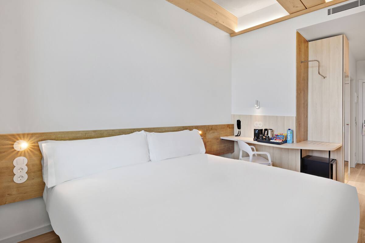 Une chambre d’hôtel moderne avec un grand lit, une tête de lit en bois, un petit bureau avec une machine à café et un miroir.