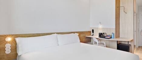 A modern hotel room with a large bed, a wooden headboard, a small desk with a coffee maker, and a mirror.