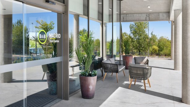 A modern hotel exterior with large windows, outdoor seating, and potted plants. - B&B Hotel Madrid Tres Cantos  (Tres Cantos)