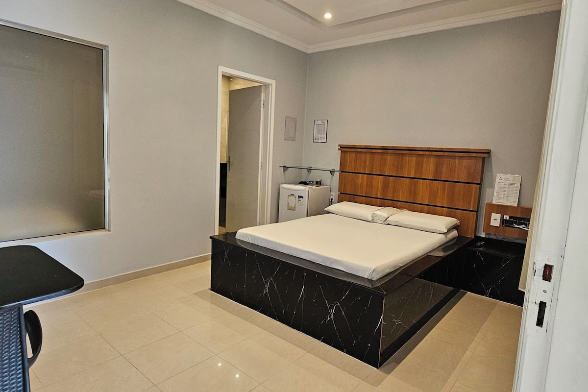 Ein Hotelzimmer mit einem Bett, einem kleinen Tisch und einem Stuhl. Ein Fenster und eine Tür zu einem weiteren Raum sind vorhanden.