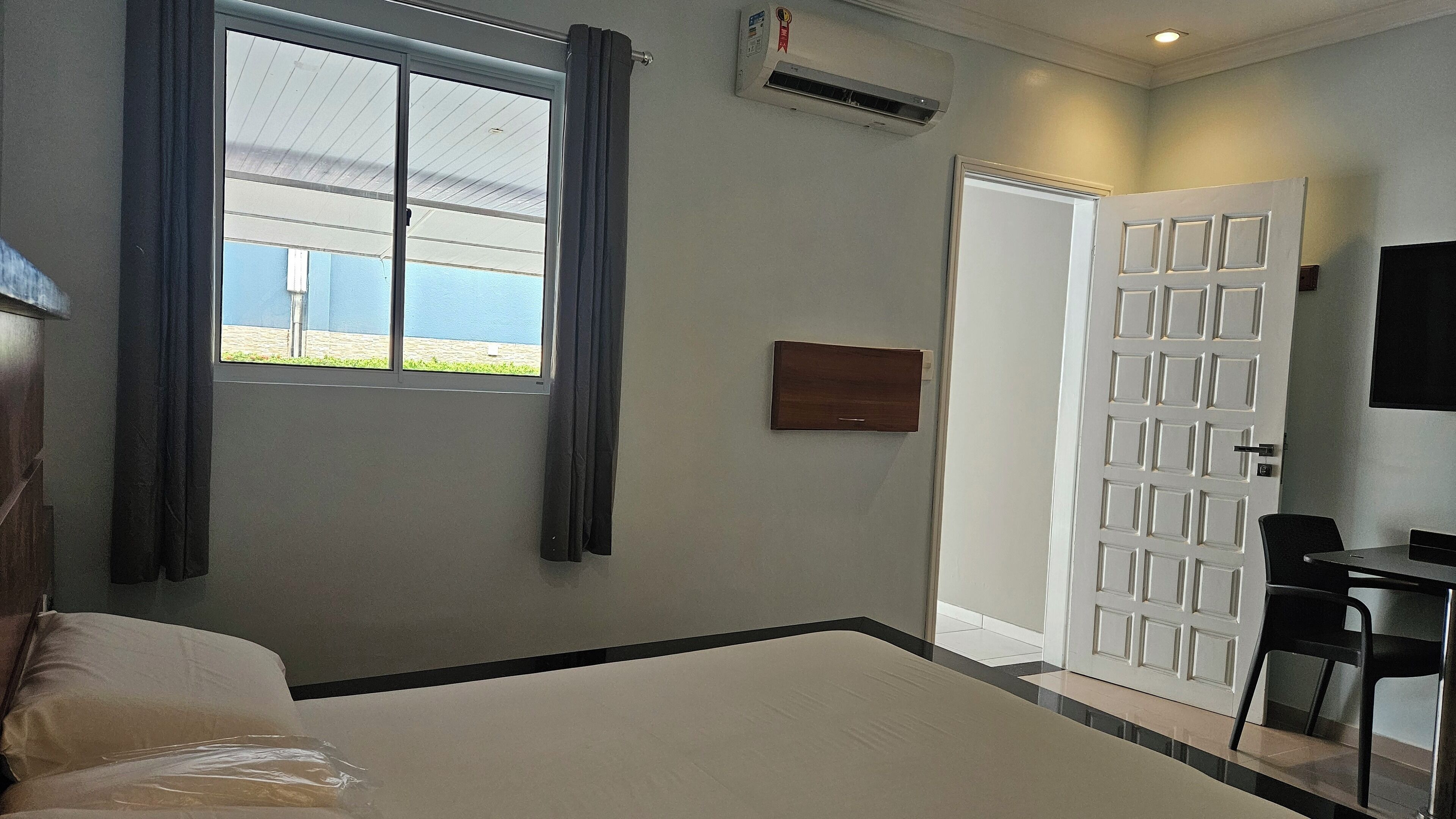A hotel room with a bed, a desk, a chair, a window with curtains, an air conditioning unit, and a door.