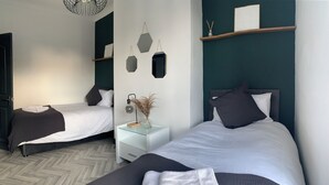 Une chambre d’hôtel moderne avec deux lits, une table de chevet blanche, une lampe fixée au mur et des miroirs hexagonaux décoratifs.