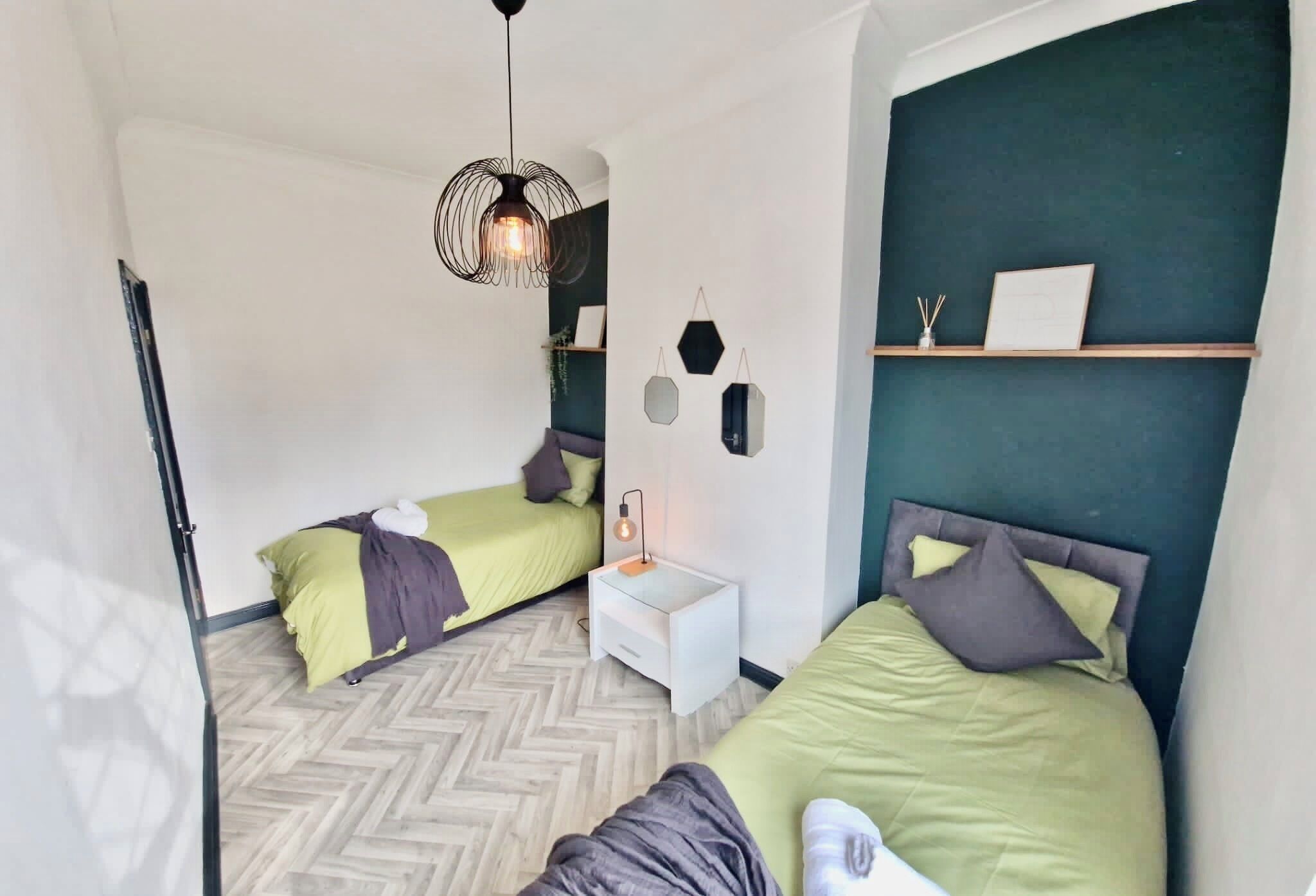Un dormitorio con dos camas, una mesita de noche blanca, una lámpara colgante y una estantería en la pared con un marco de fotos y adornos.