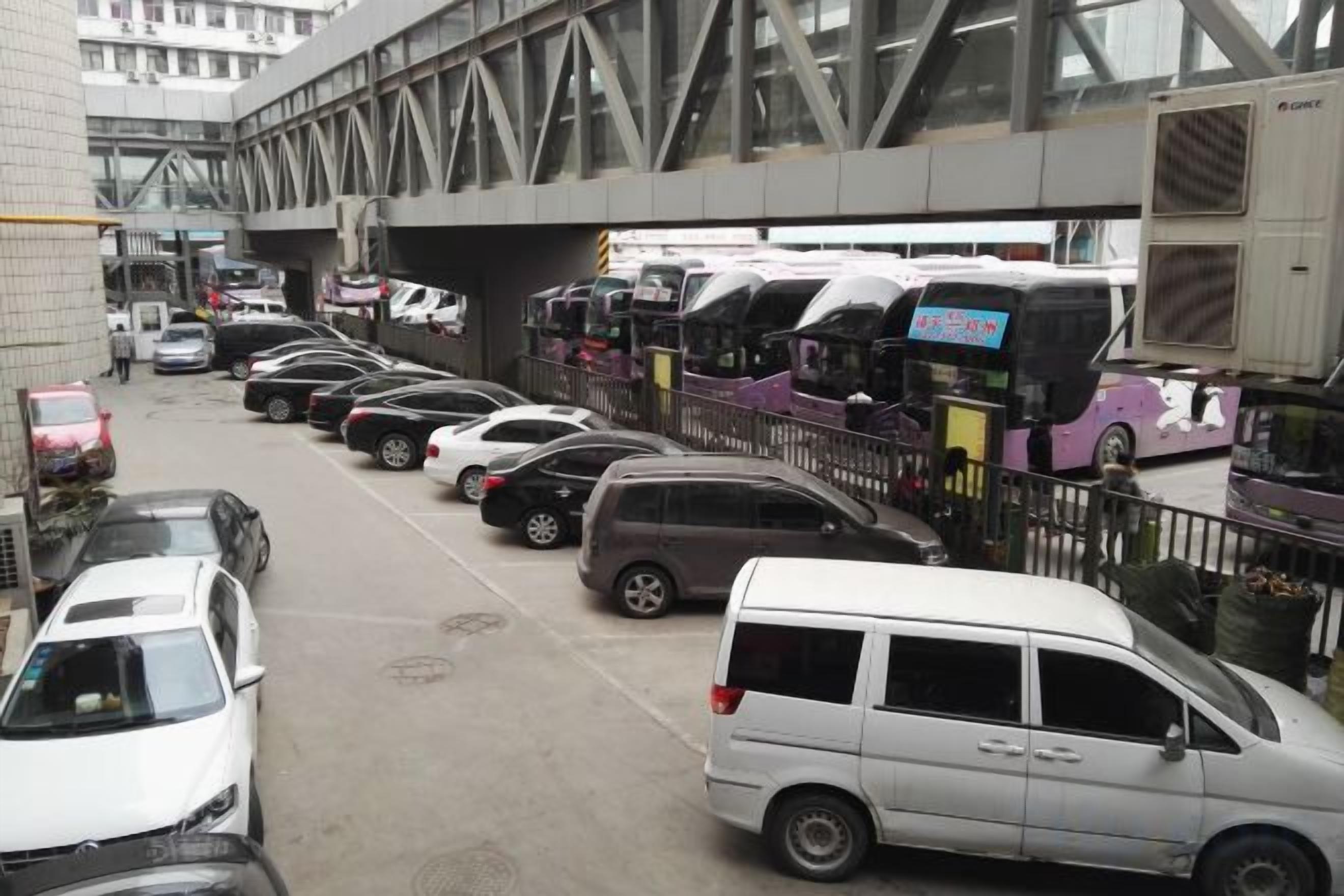 Un estacionamiento con varios coches y autobuses, una acera peatonal y una estructura de puente al fondo.