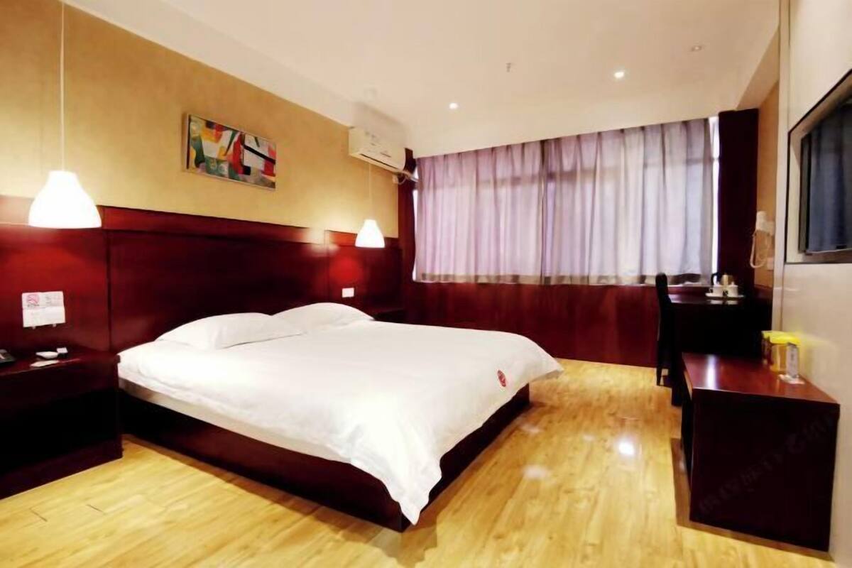 A hotel room with a wooden floor, a large bed with white linens, a desk with a chair, and a window with curtains.