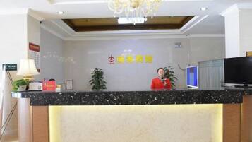 Ein Hotelrezeptionstresen mit Rezeptionistin, einer Chandelierleuchtre, einem digitalen Bildschirm und einem Schild mit chinesischen Schriftzeichen.