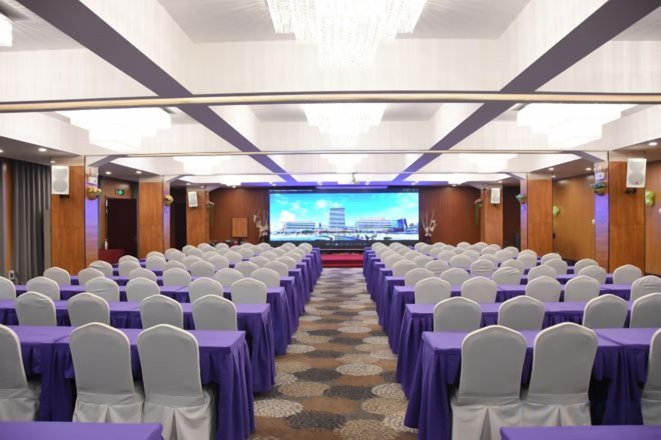 A conference room with rows of tables and chairs, a stage with a large screen, and a carpeted floor.