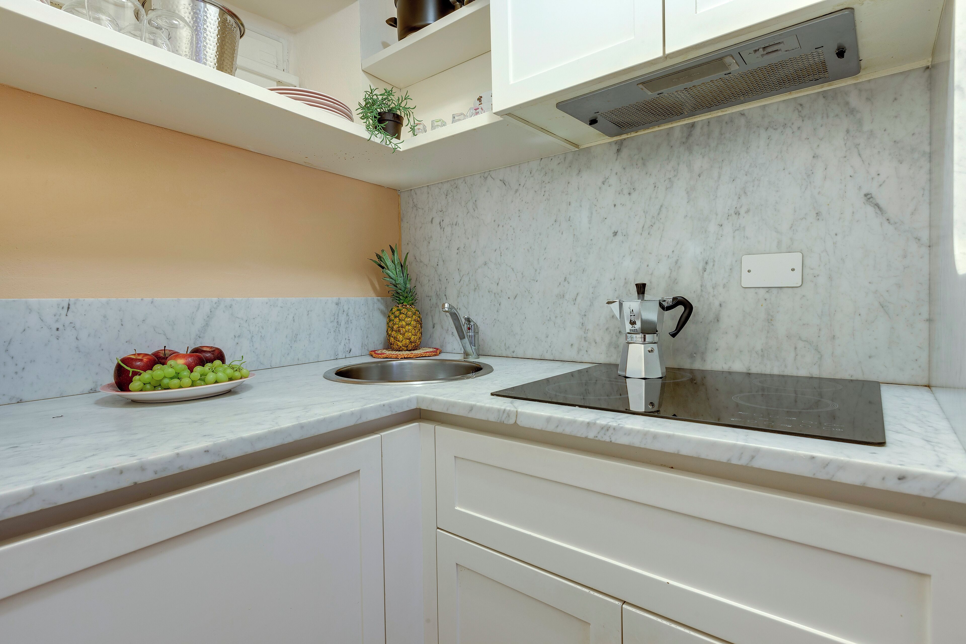 Una cocina con armarios blancos, encimera de mármol, pileta y campana extractora de acero inoxidable.