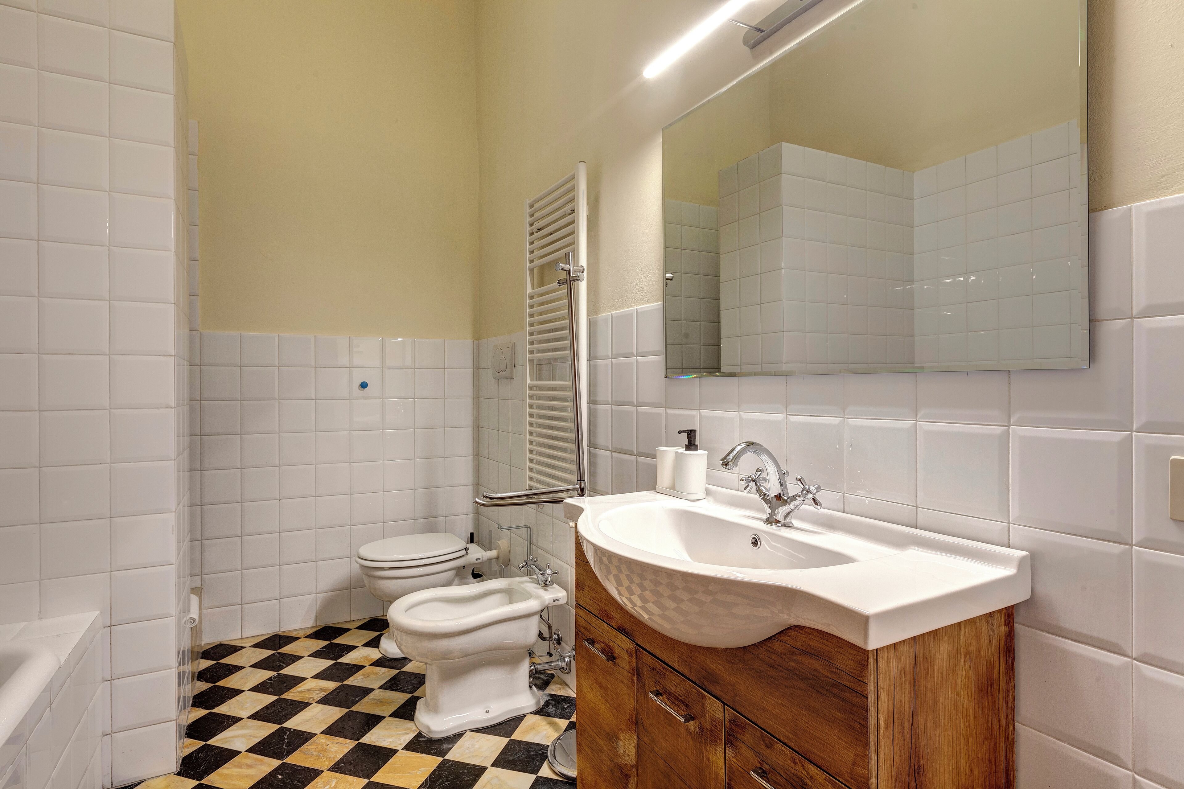 Une salle de bain avec des murs recouverts de carreaux blancs, un sol à carreaux, un lavabo blanc et un miroir au-dessus du lavabo.