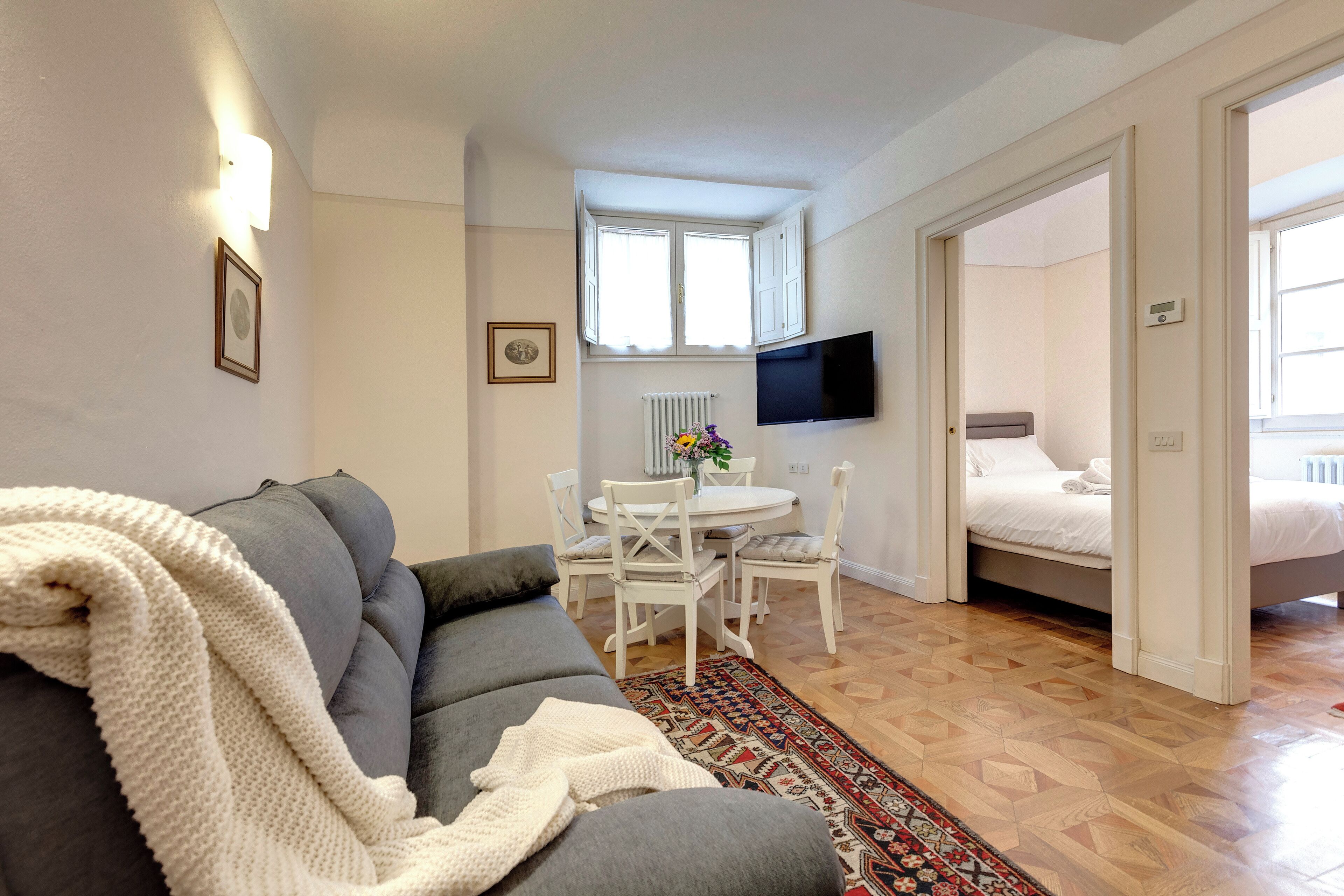 Een moderne woonkamer met een grijze bank, een witte gebreide plaid, een kamerkleed met een patroon, een eettafel met stoelen, een tv die aan de muur is bevestigd en een bed in de aangrenzende kamer.