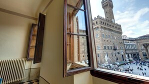 Vista desde una ventana sobre un edificio histórico con una torre de reloj, una fuente y una plaza concurrida.