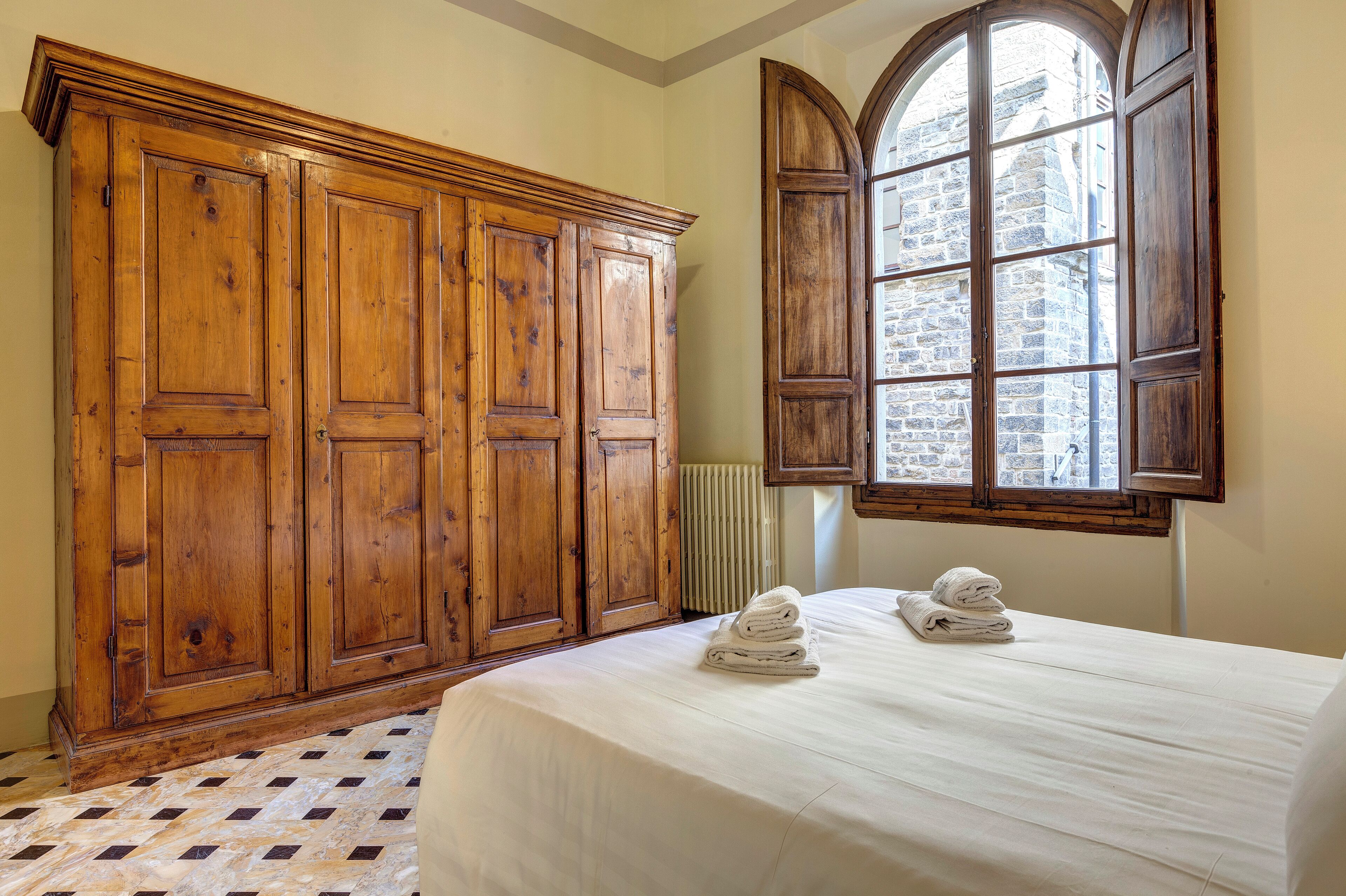 Un dormitorio con un armario grande de madera, una cama con ropa blanca y una ventana que ofrece vistas a un edificio de piedra.