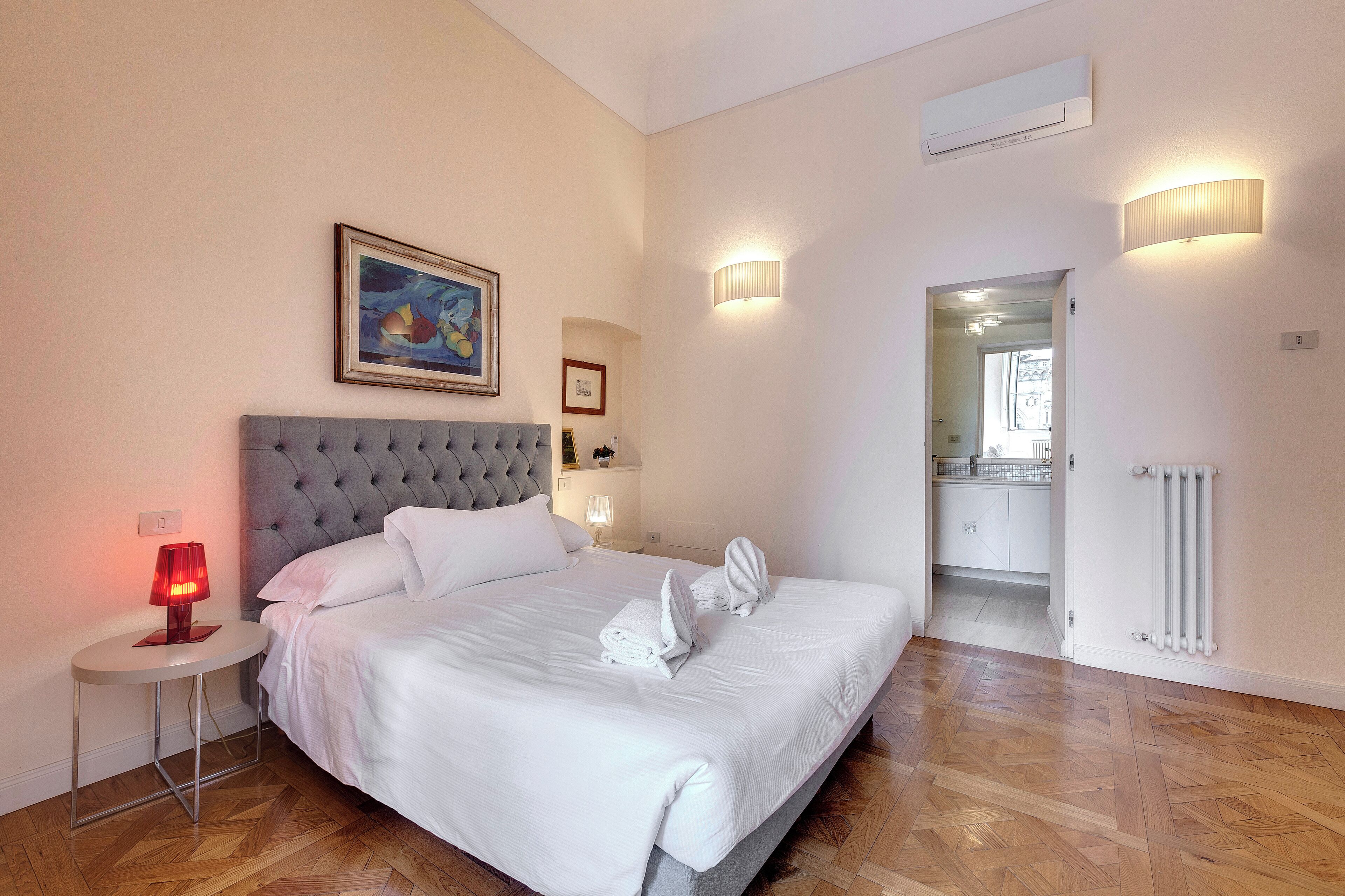 A neatly made bed with white linens, a round bedside table with a red lamp, and a framed picture on the wall.
