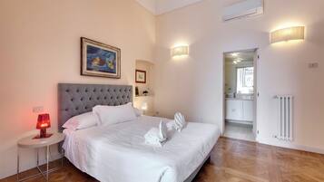 Een netjes opgemaakt bed met witte lakens, een rond nachtkastje met een rode lamp en een lijst met een afbeelding aan de muur.